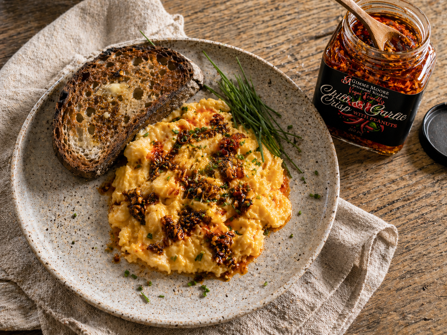 Soft scrambled eggs finished with a spoonful of Gimme Moore Chilli & Garlic Crisp on a speckled cream plate, sourdough toast, fresh chives, the open jar to one side